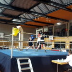 Zwei Boxer im Ring. Davor (sitzend) der Ringrichter